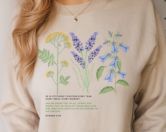 May include: Beige sweatshirt featuring embroidered floral designs in yellow, purple, and blue. The text reads "HE IS STITCHING TOGETHER EVERY TEAR, EVERY SMILE, EVERY MOMENT." and "ROMANS 8:28". A comfortable, casual top.