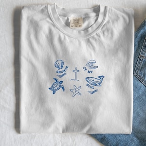Puede incluir: Camiseta blanca con ilustraciones azules de una concha, tortuga, cruz, estrella de mar, ola y ballena. El texto "ANCHOR" y "OF MY SOUL" está impreso. La camiseta está doblada.