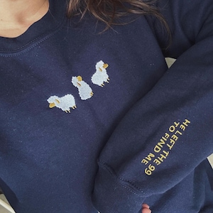Könnte beinhalten: Ein marineblauer Pullover mit einem weißen gestickten Design aus drei Schafen. Der Text "HE LEFT THE EGG TO FIND ME" ist in Gelb auf dem Ärmel gestickt.