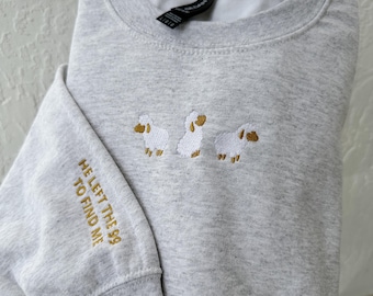 May include: A light gray heather crewneck sweatshirt featuring an embroidered design of three white sheep with gold-colored faces and legs. The sleeve is embroidered with the gold-colored text HE LEFT THE 99 TO FIND ME in a sans-serif font.