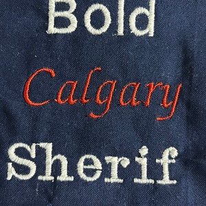 May include: White embroidered text on a dark blue fabric background reads "Bold Calgary Sheriff".