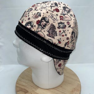 May include: A beige welding cap with a black trim and a pattern of pin-up girls, skulls, anchors, and ships.
