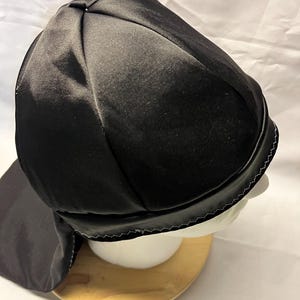 Pode incluir: Durag de cetim preto com aba e borda costurada. O durag é exibido em uma cabeça de manequim branca e um suporte de madeira. O tecido tem um leve brilho.