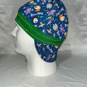 May include: A blue welding cap featuring a repeating pattern of cartoon characters, planets, and spaceships. The cap has a green trim and the words "Rick and Morty" are printed on the fabric. The cap is displayed on a white mannequin head.