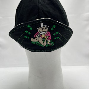 May include: Black cycling cap with a green, white, and pink embroidered design of a cartoon character sitting on a pile of money.