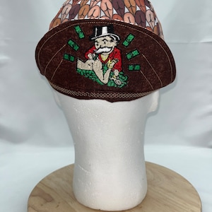 May include: A brown and tan welding cap with a unique design. The top of the cap features a pattern of brown buttocks. The front of the cap has an embroidered image of a Monopoly man surrounded by money.