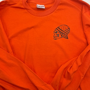 Puede incluir: Camiseta de manga larga naranja con un gráfico de calavera negra que lleva un pañuelo con estrellas y rayas. La calavera mira hacia la izquierda. La camiseta tiene cuello redondo.