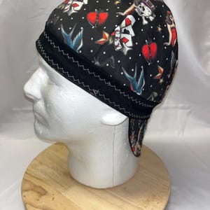 May include: Black skull cap with a repeating pattern of tattoo-style imagery. The design includes hearts, playing cards, swallows, and pin-up girls. The cap has a black trim with white stitching.