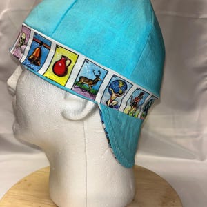 May include: A light blue welding cap with a patterned border featuring colorful illustrations. The cap is displayed on a white mannequin head, showcasing its design and fit. The border includes images of a bell, a deer, and other graphics.