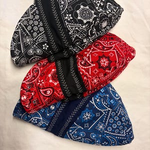 May include: Three folded fabric caps featuring a traditional paisley bandana print in black, red, and blue. Each cap has a solid black or dark blue fabric band across the center with decorative white zigzag stitching along the edges.