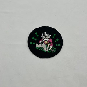 Puede incluir: Un parche bordado negro y verde con un personaje de dibujos animados sentado sobre una pila de dinero. El personaje lleva una chaqueta rosa, un sombrero blanco y un bigote. El parche está rodeado de billetes verdes.