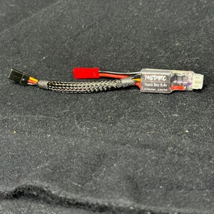 May include: A small electronic device with the text "NSDFC Nano Bec 8.4v" printed on it. The device has a black and silver braided cable, a red connector, and a black connector. The device is on a black surface.