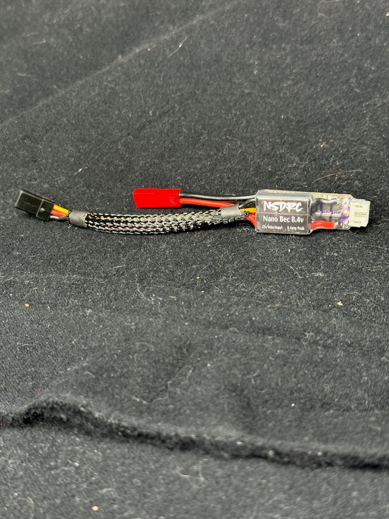 May include: A small electronic device with the text "NSDFC Nano Bec 8.4v" printed on it. The device has a black and silver braided cable, a red connector, and a black connector. The device is on a black surface.