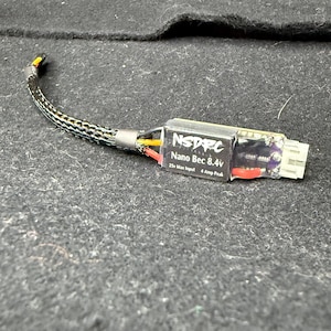 May include: A black and gray electronic component labeled "NSDRC Nano Bec 8.4v" with a white connector and a black and gray braided wire. The component has red, yellow, and black wires visible.