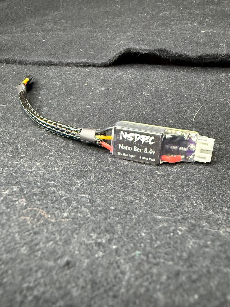 May include: A black and gray electronic component labeled "NSDRC Nano Bec 8.4v" with a white connector and a black and gray braided wire. The component has red, yellow, and black wires visible.