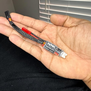 May include: A black and red electronic component with the text "NSDPC Nano Bec 8.4v" printed on a white label. The component has a black braided cable and is held in a hand.