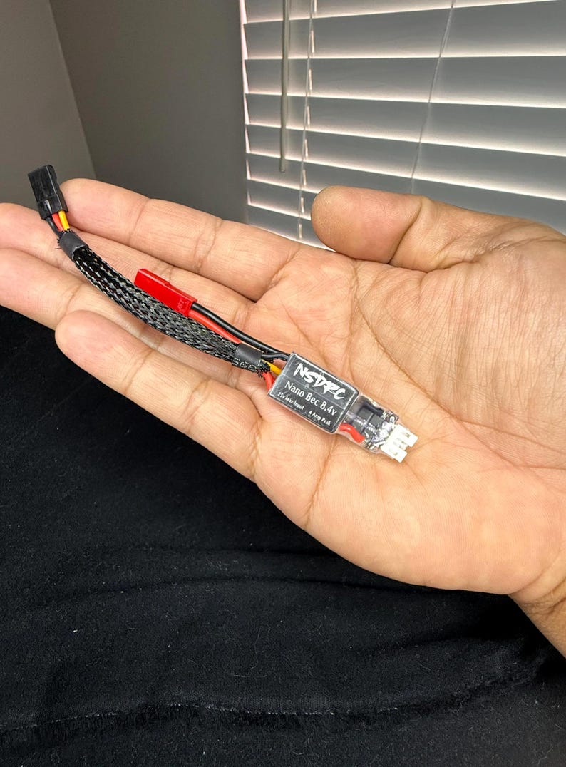 May include: A black and red electronic component with the text "NSDPC Nano Bec 8.4v" printed on a white label. The component has a black braided cable and is held in a hand.