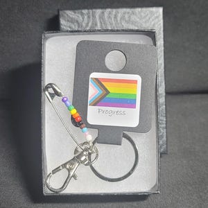 May include: A keychain with a rainbow pride flag charm, a safety pin, and a beaded chain. The charm has the text "Progress" on it.