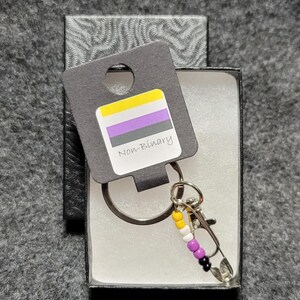 May include: A keychain with a silver ring, a silver clasp, and a beaded charm. The beads are yellow, white, purple, and black, representing the non-binary pride flag. A tag attached to the ring reads "Non-Binary" with the non-binary pride flag printed on it.