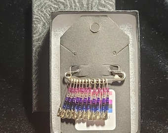 May include: A safety pin brooch with a beaded design in the colors of the bisexual pride flag. The brooch is displayed in a black box with a white tag.