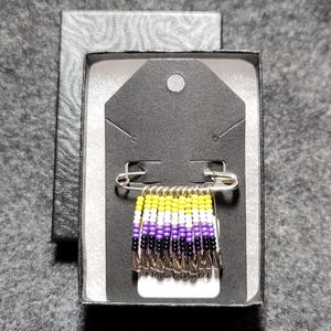 Peut inclure: Une épingle à nourrice de fierté non-binaire avec des perles jaunes, blanches, violettes et noires. L'épingle est fixée à une épingle de sûreté et est présentée dans une boîte noire avec une étiquette blanche qui dit "Non-binaire".