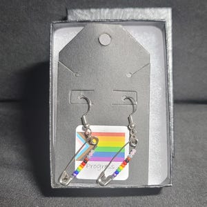 May include: A pair of silver safety pin earrings with rainbow colored beads. The earrings are in a black box with a progress pride flag and the word "Progress" printed on it.
