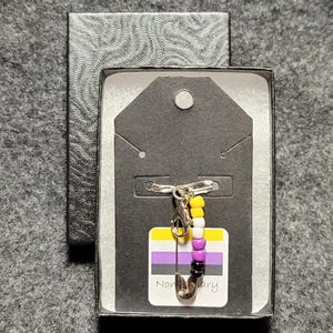 Może przedstawiać: Srebrna szpilka bezpieczeństwa z koralikową zawieszką w kolorach flagi dumy osób niebinarnych. Zawieszka jest przymocowana do kartki z tekstem "Niebinarny".