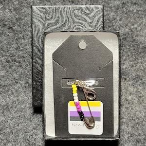 May include: A non-binary pride flag charm with a safety pin attached to a silver keychain. The charm is made of small beads in the colors of the non-binary pride flag: purple, white, yellow, black, and yellow. The charm is in a black box with a white tag that says "Non-Binary".