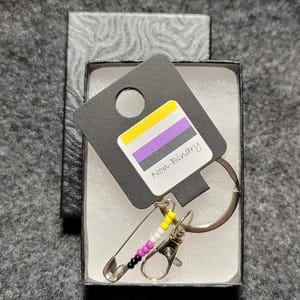Pode incluir: Um chaveiro de orgulho não binário com um alfinete de segurança e um amuleto de contas. O amuleto é feito de contas amarelas, brancas, roxas e pretas. O chaveiro está em uma caixa preta com uma etiqueta que diz "Não binário".