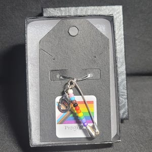 Puede incluir: Un imperdible de plata con un abalorio de cuentas de arcoíris y una tarjeta de bandera del orgullo progresista. La tarjeta dice "Progress Pride".