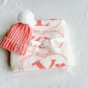 Puede incluir: Un gorro de bebé de punto rosa con un pompón blanco y una manta de punto blanco con un diseño rosa. La manta tiene una cinta blanca atada alrededor.