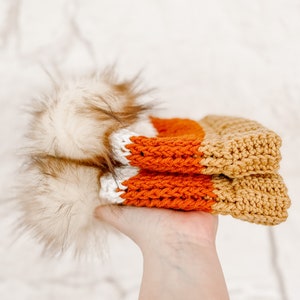 Puede incluir: Un gorro de punto con un patr&oacute;n de rayas blancas, naranjas y marrones. El gorro tiene un pomp&oacute;n esponjoso en la parte superior.