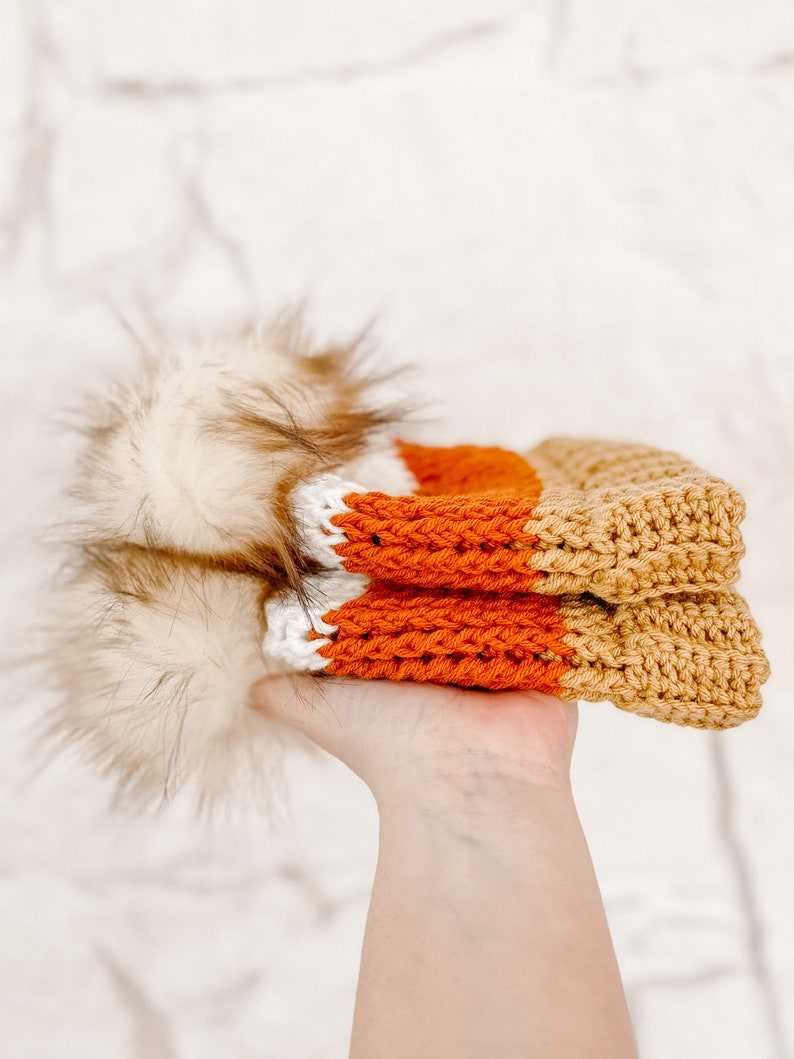 Puede incluir: Un gorro de punto con un patr&oacute;n de rayas blancas, naranjas y marrones. El gorro tiene un pomp&oacute;n esponjoso en la parte superior.