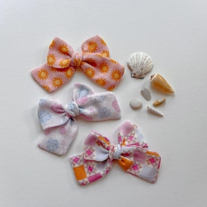 May include: Three fabric hair bows in pink, white, and orange patterns. One bow features a sunflower design, another has a floral pattern, and the third has a geometric design. Seashells and small stones are arranged nearby on a white surface.