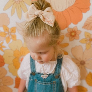 Puede incluir: Una ni&ntilde;a peque&ntilde;a con cabello rubio que lleva una camisa blanca y un mono azul. Tiene un lazo beige con la palabra "Lola" bordada en el pelo. Lleva un collar con un colgante de mariposa.