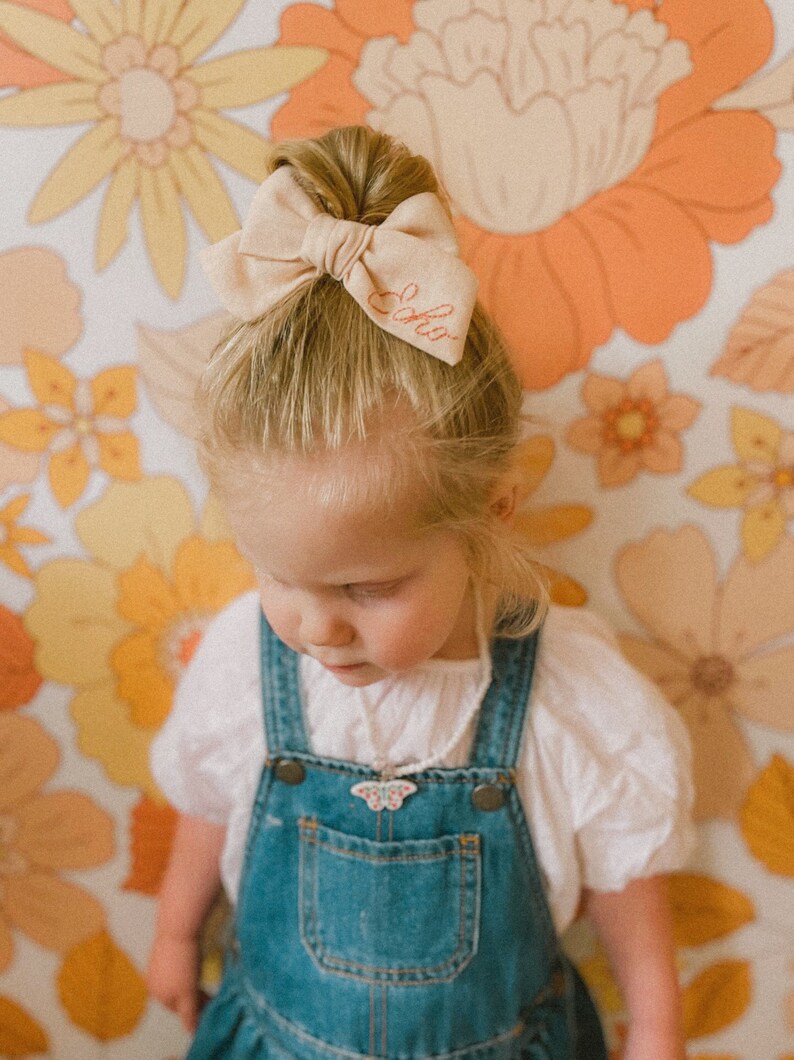 Puede incluir: Una ni&ntilde;a peque&ntilde;a con cabello rubio que lleva una camisa blanca y un mono azul. Tiene un lazo beige con la palabra "Lola" bordada en el pelo. Lleva un collar con un colgante de mariposa.