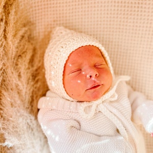 May include: A newborn baby wearing a white crocheted hat and matching sweater. The baby is sleeping peacefully on a soft, beige blanket.