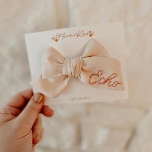 Puede incluir: Un lazo de tela beige con la palabra "Echo" bordada en hilo naranja. El lazo est&aacute; sobre una tarjeta blanca con el texto "Flora + Roo" en cursiva marr&oacute;n.