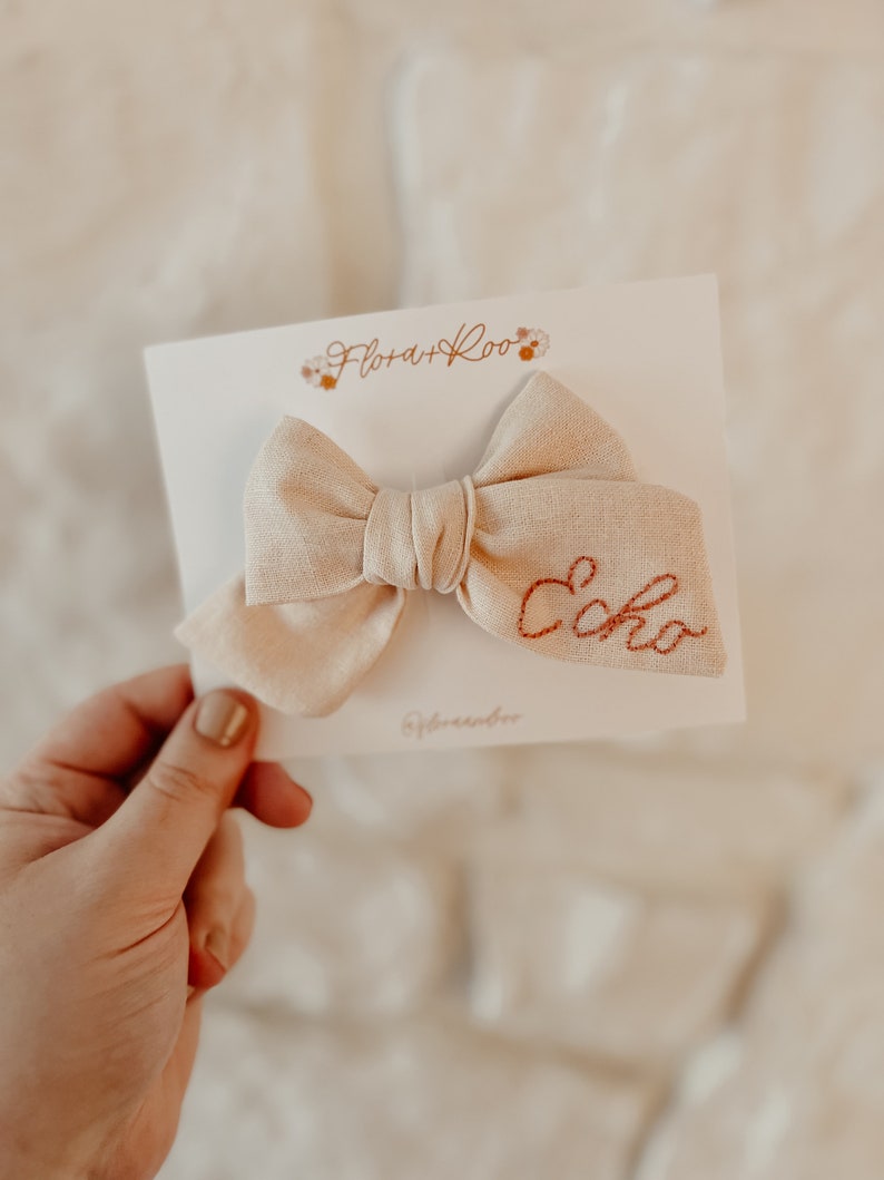 Puede incluir: Un lazo de tela beige con la palabra "Echo" bordada en hilo naranja. El lazo est&aacute; sobre una tarjeta blanca con el texto "Flora + Roo" en cursiva marr&oacute;n.