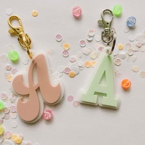 May include: Two acrylic letter A keychains with metal lobster claw clasps. One letter is a dusty rose color in a cursive font, and the other is a light mint green in a block font. Both are layered over a clear acrylic base and surrounded by small colorful beads.