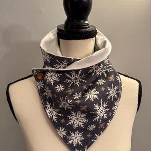 May include: A dark blue and white snowflake patterned neck warmer with a white fleece lining. The neck warmer is secured with a wooden button. The design features white and gold snowflakes.