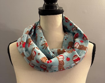 May include: A light blue infinity scarf with a pattern of brown sloths wearing red Santa hats and holding ornaments. The sloths are on a white background with green pine branches and red berries.