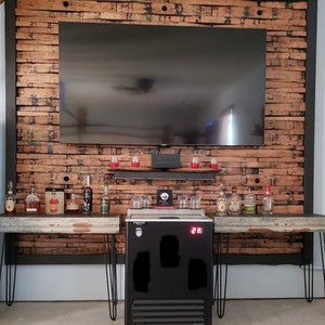 K&ouml;nnte beinhalten: Ein schwarzer K&uuml;hlschrank mit einem digitalen Temperaturdisplay, das auf 21 Grad Fahrenheit eingestellt ist. Der K&uuml;hlschrank steht auf einem Holztisch mit einem schwarzen Metallrahmen. Auf dem Tisch stehen Flaschen mit Spirituosen und Gl&auml;ser. Der K&uuml;hlschrank steht vor einer Wand aus wiederverwendeten Holzbrettern.