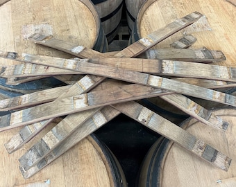 May include: A pile of wooden barrel staves, some with metal bands, arranged in a criss-cross pattern. The staves are a light brown color and have a rough, textured surface.
