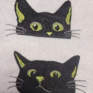 以下が含まれることがあります： 白い背景に、黄色のアクセントが付いた黒い猫の顔の刺繍。猫の目と内側の耳は黄色で、ひげが見えます。このデザインはおそらく装飾品またはテキスタイル用です。