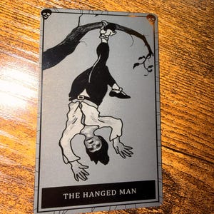 May include: A tarot card featuring an illustration of a figure hanging upside down from a tree branch. The figure is dressed in black pants, a white shirt, and black shoes. The card is labeled "THE HANGED MAN" in a black box.