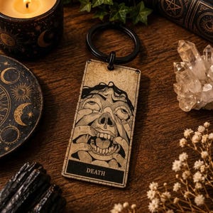 May include: A rectangular keyring featuring a beige tarot card design with a black ink illustration of a face with hands pulling at the skin. The word DEATH is printed in a black rectangular box at the bottom. It has a black metal ring.
