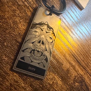 Könnte beinhalten: Ein rechteckiger Schlüsselanhänger mit einem schwarzen Metallring. Der Schlüsselanhänger zeigt eine detaillierte Schwarz-Weiß-Illustration eines Gesichts mit Händen, die Augen und Mund bedecken. Das Wort "DEATH" ist in einem schwarzen Rechteck unten aufgedruckt.