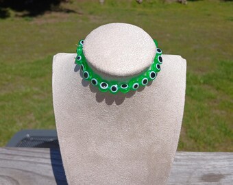 May include: A green evil eye bracelet with white pupils. The bracelet is on a white display stand.