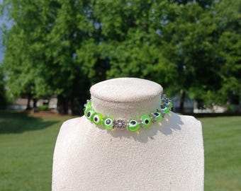 May include: A green evil eye choker necklace with silver flower accents. The necklace is on a white mannequin bust.